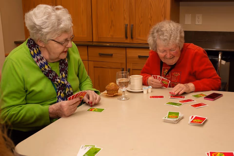 Two elderly women sitting at a table playing a card game. One woman is wearing a green sweater and a colorful scarf, while the other is wearing a red sweater. On the table are a glass of water, a coffee cup with creamers, a muffin, and a smartphone. The background shows wooden cabinets and a countertop.