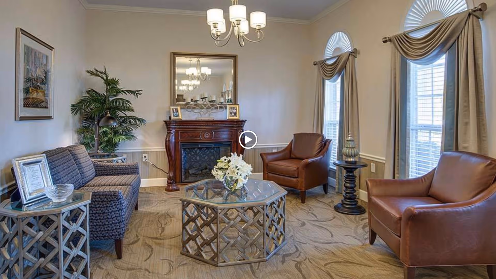 A cozy senior living facility common area with a patterned sofa, two brown leather armchairs, a glass-top coffee table, a wooden fireplace with a mirror above it, two large windows with beige curtains, a side table with a decorative lamp, a potted plant, and framed artwork on the wall.