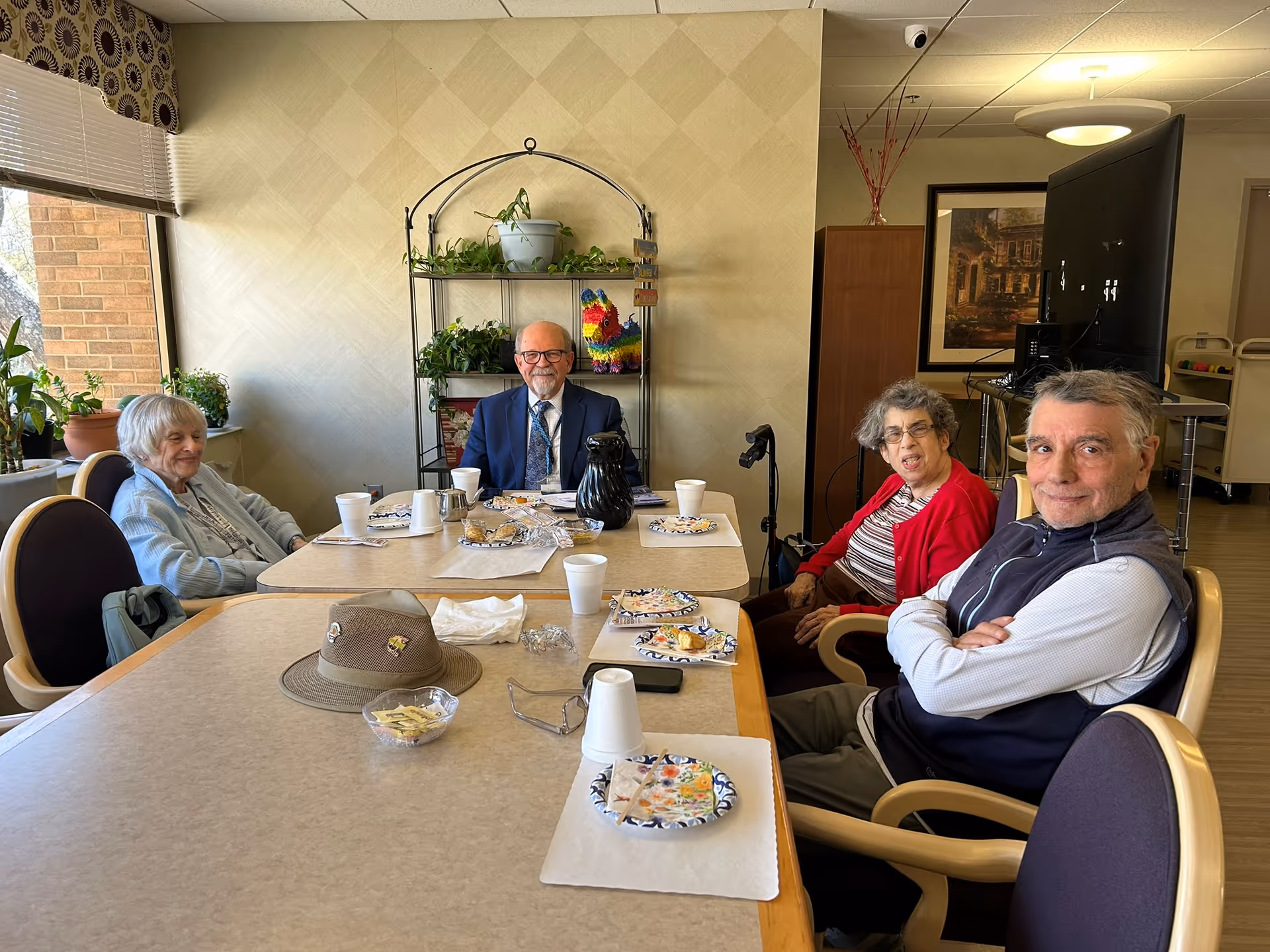 Four elderly people sitting around a dining table in a well-lit room with large windows and plants on the windowsill. The table has paper plates with food remnants, disposable cups, a hat, and some small items. Behind them is a metal shelf with potted plants and a colorful decorative item. The room has light-colored walls and wooden flooring.