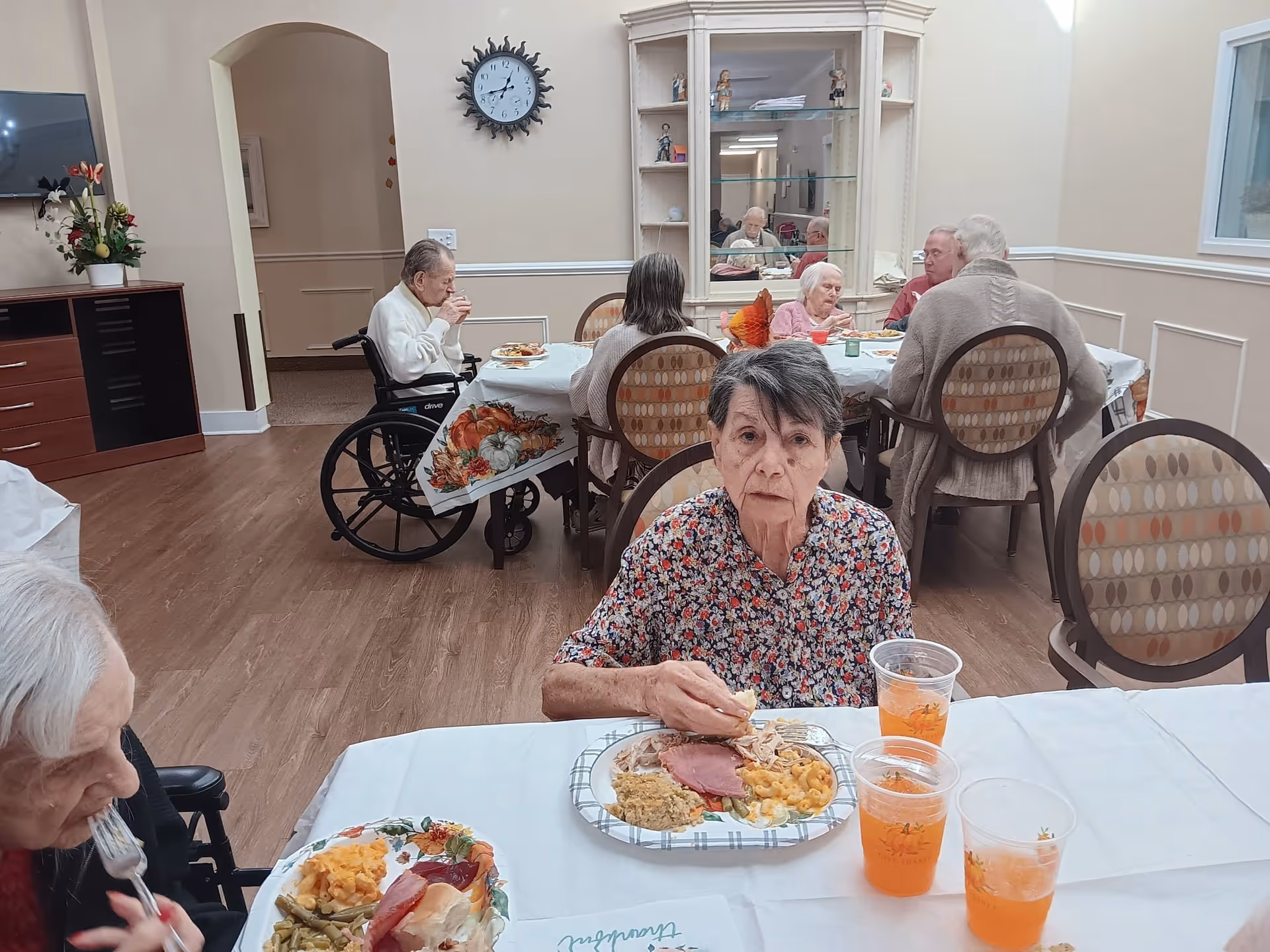 A group of elderly people seated around tables in a dining room at an assisted living facility. They are eating a meal that includes ham, macaroni and cheese, green beans, and other side dishes. The room has light-colored walls, a clock on the wall, a cabinet with decorative items, and a television mounted on the wall.