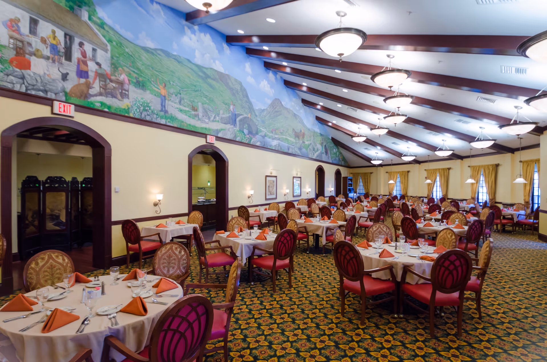 A spacious dining room with multiple round tables covered with white tablecloths and set with orange folded napkins, glasses, and silverware. The room features patterned carpet, upholstered chairs with red and gold designs, large windows with yellow curtains, and a high ceiling with exposed wooden beams and hanging light fixtures. A large mural depicting a scenic landscape with people and animals decorates the upper part of one wall.