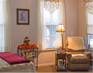A cozy living room area with a beige recliner chair draped with a cream-colored knitted blanket, a wooden side table with a lamp and small decorative items, two windows with floral valances letting in natural light, a small table decorated with autumn-themed items and framed photos, and a bed with a red blanket partially visible on the left side.