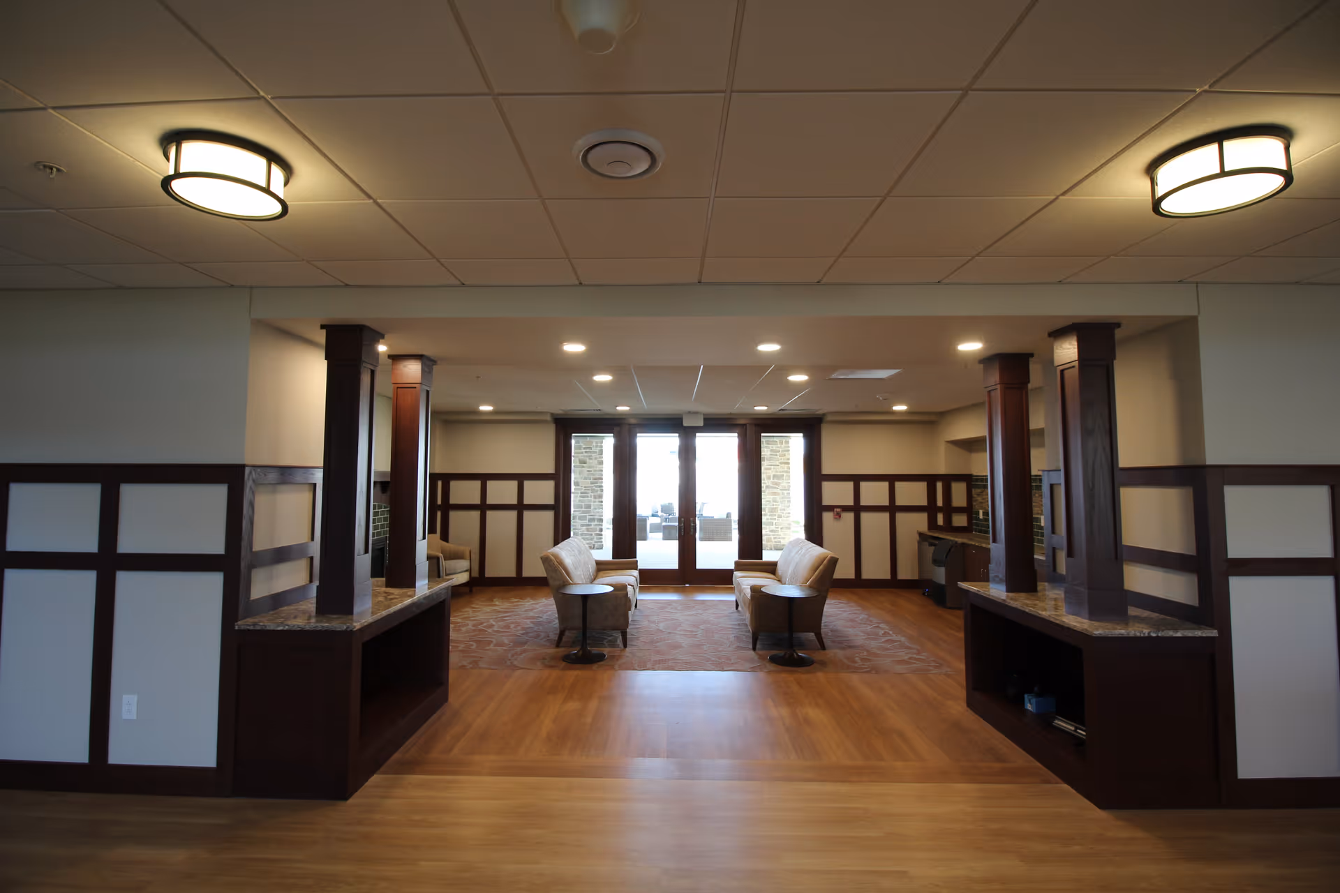 A spacious interior common area with wooden flooring and beige walls featuring dark wood paneling. The room has two ceiling lights and recessed lighting. There are two pairs of beige upholstered chairs facing each other with small round tables between them. At the far end, there are large glass doors leading outside, allowing natural light to enter the room.