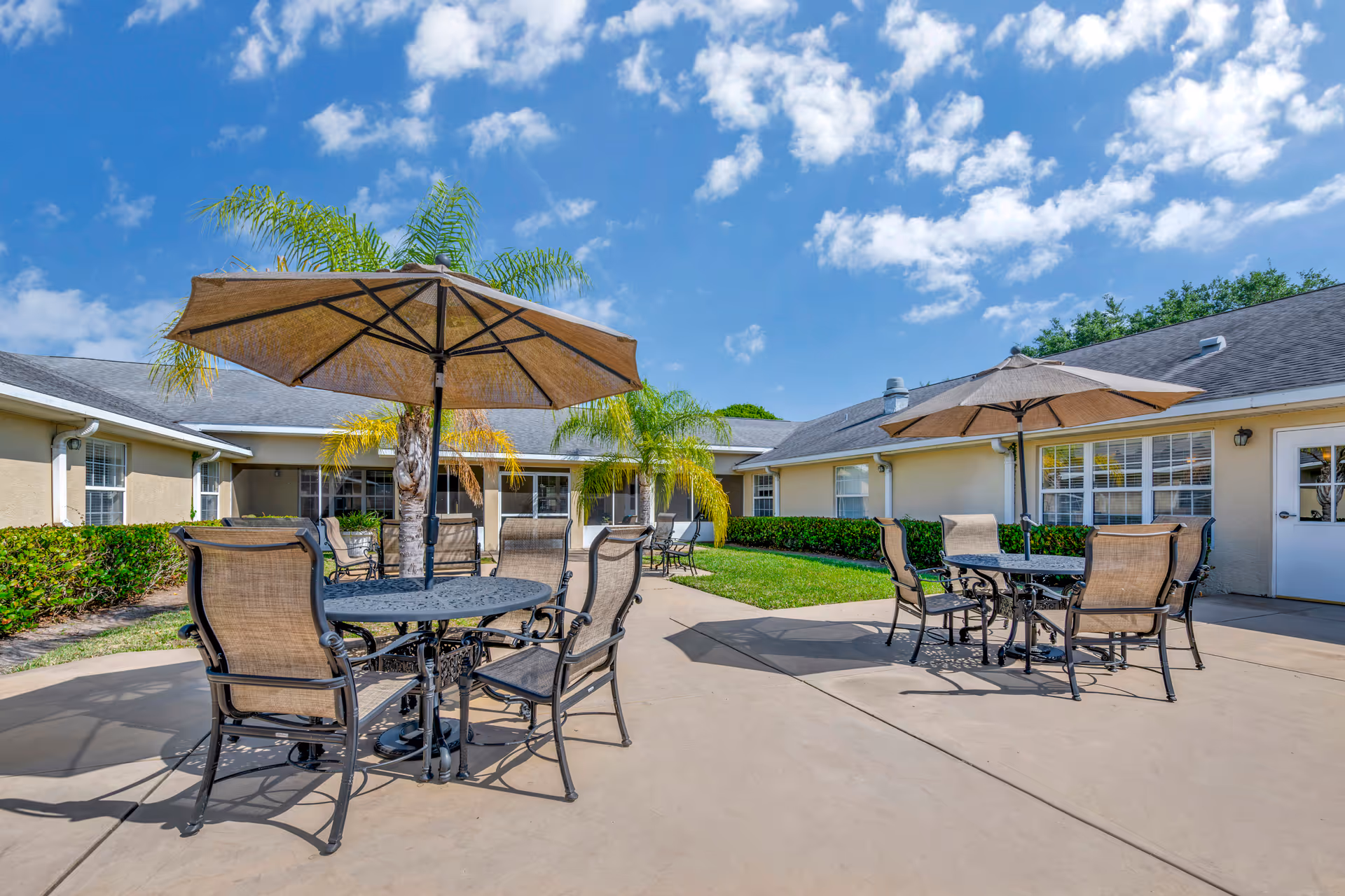 Outdoor patio area at Brookdale Vero Beach South Memory Care with two round metal tables, each surrounded by four mesh chairs and shaded by large beige umbrellas. The patio is paved and bordered by green grass, palm trees, and beige buildings under a blue sky with scattered clouds.