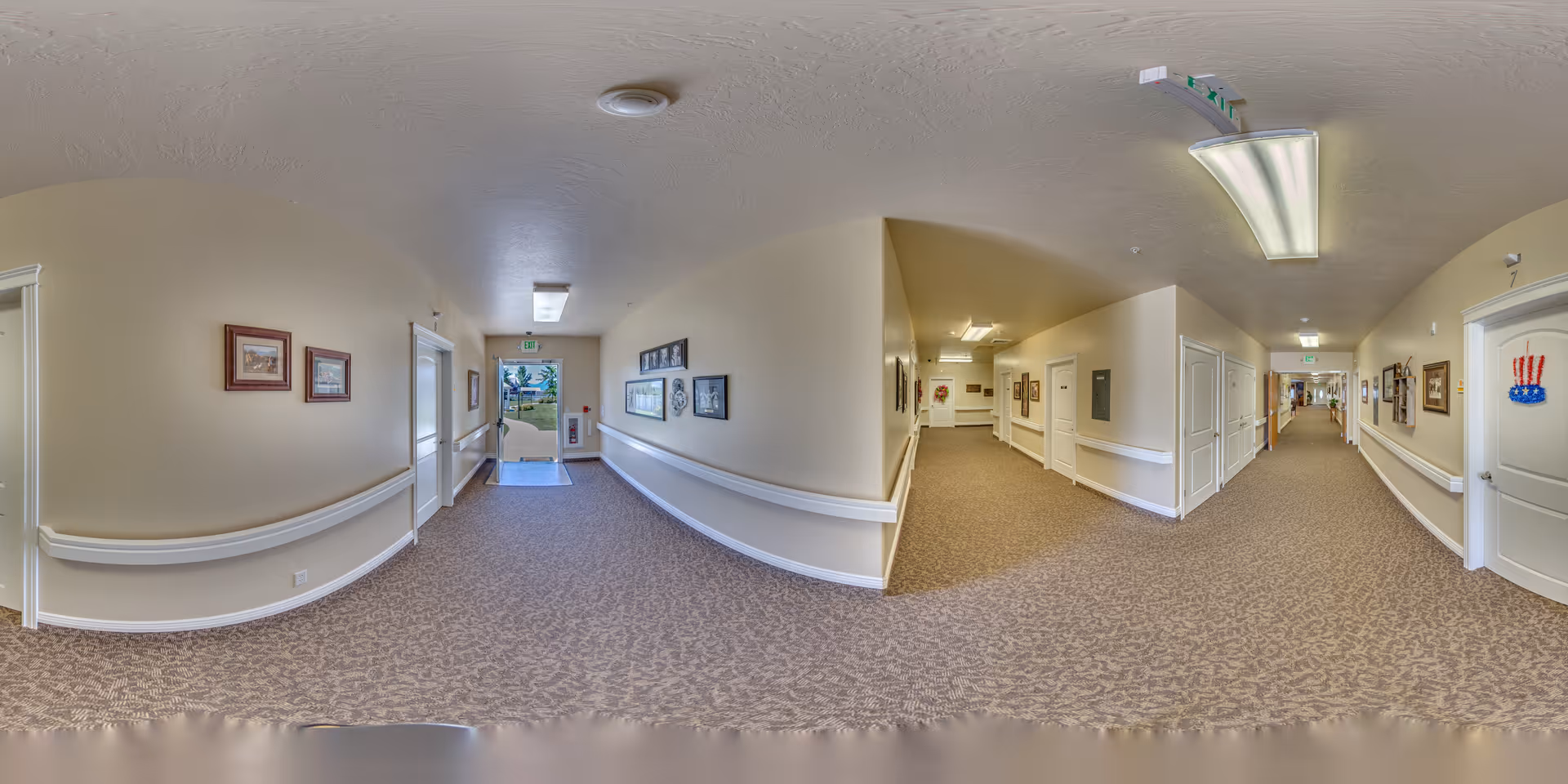 Wide-angle view of a clean, well-lit hallway in a senior living facility with beige walls, carpeted floors, handrails along the walls, framed pictures, and exit doors at the end.