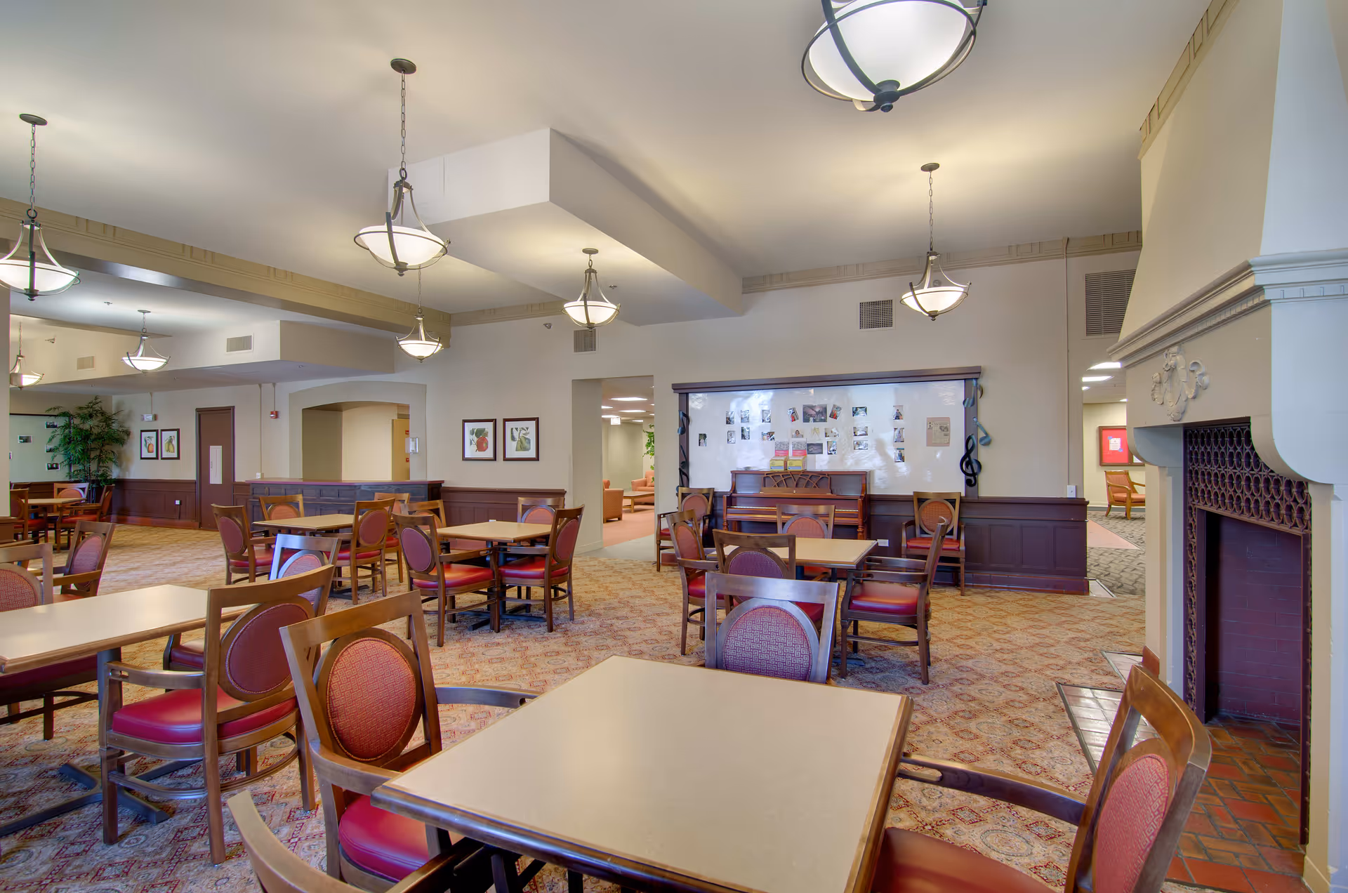 A spacious dining room in a senior living facility with multiple square tables and wooden chairs with red cushioned seats. The room features a carpeted floor with a patterned design, several hanging light fixtures, framed artwork on the walls, a piano against one wall with photos and notes displayed above it, and a fireplace on the right side.
