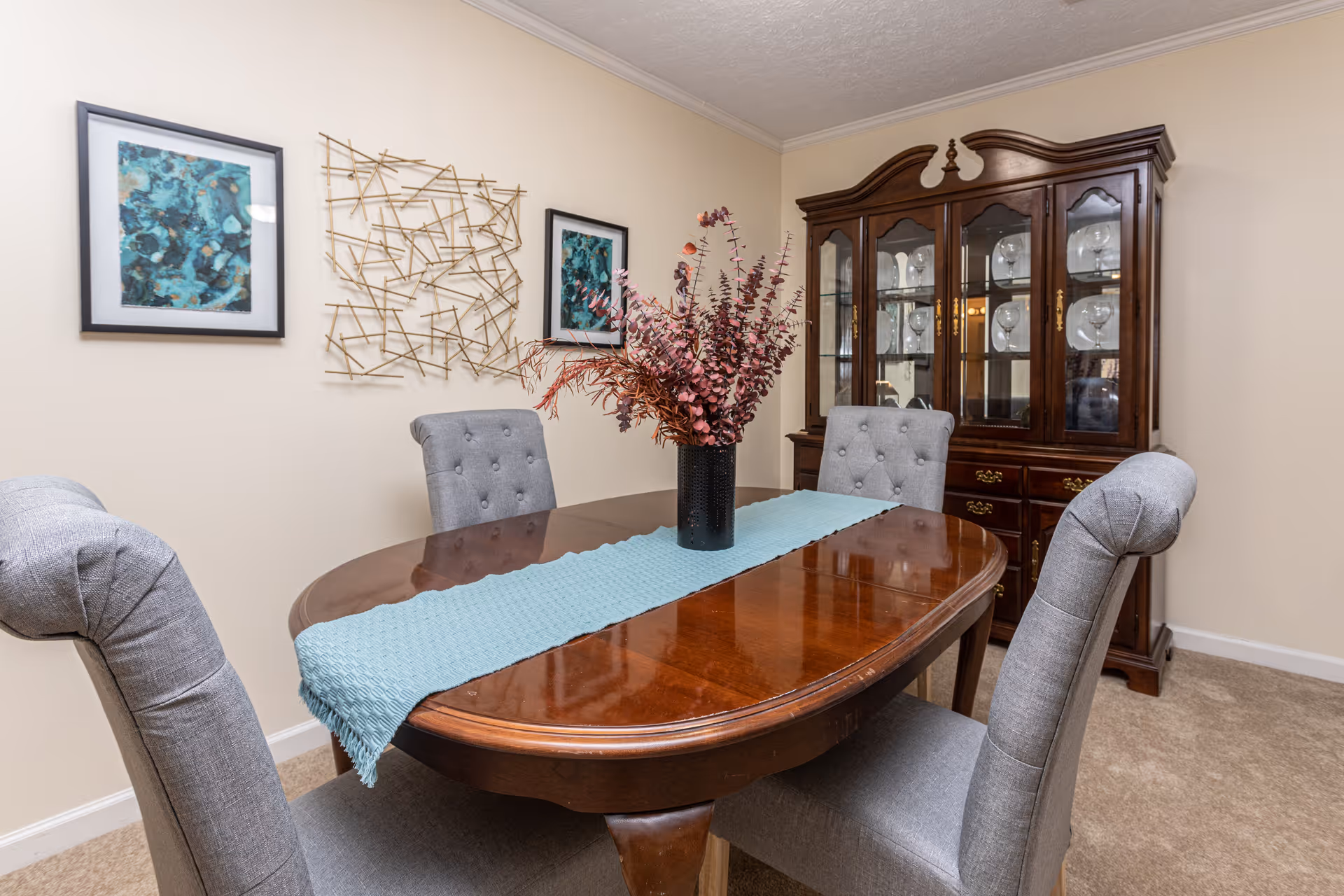Dining room with a polished wooden table, four gray upholstered chairs, a floral centerpiece, wall art, and a china cabinet.