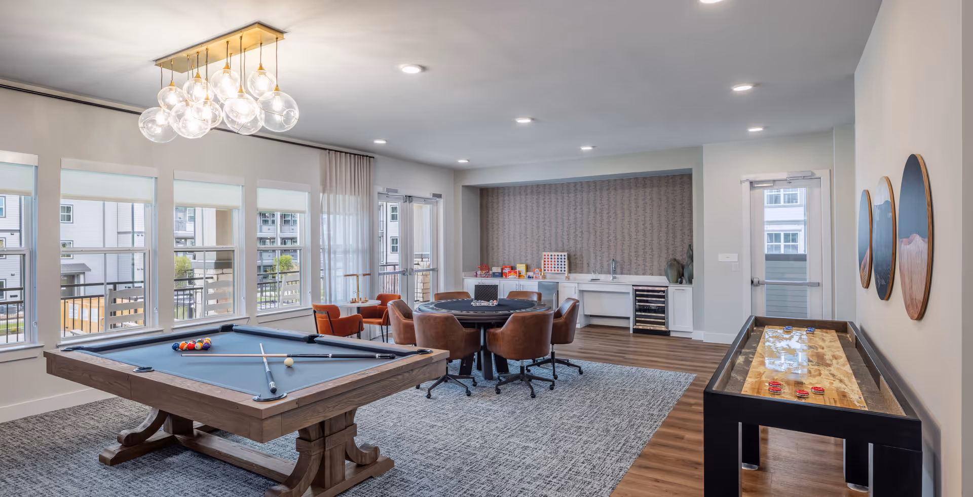 A bright and modern game room with a pool table, a round table with chairs, a shuffleboard table, and a small kitchenette area with snacks and a wine cooler. Large windows and glass doors let in natural light, and a contemporary chandelier hangs from the ceiling.