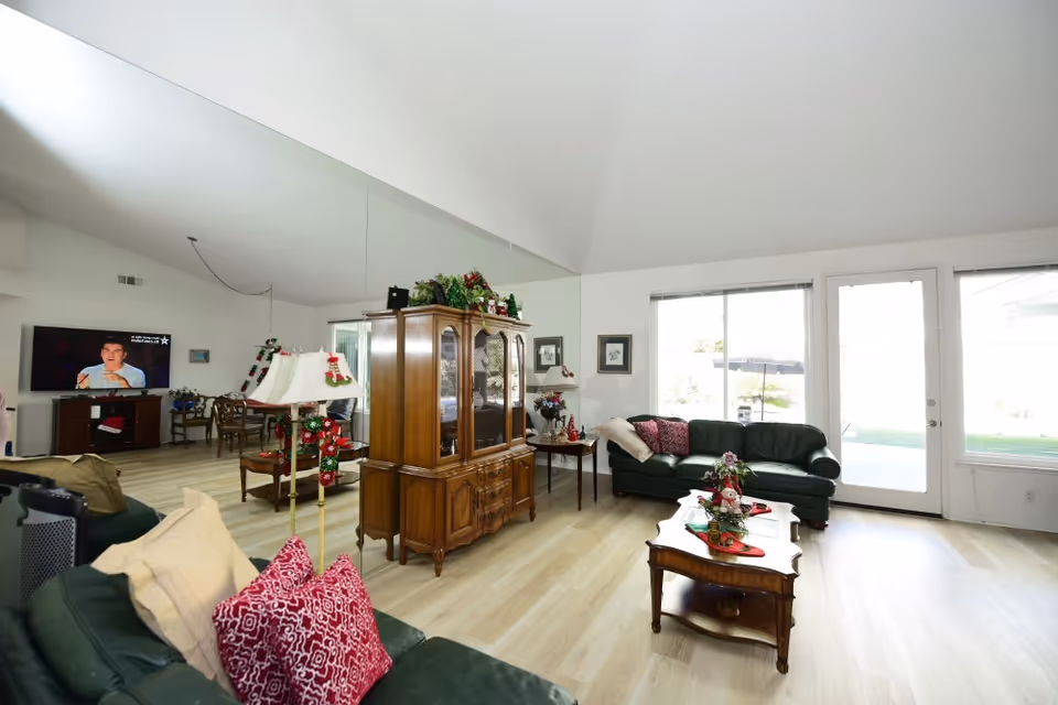 Spacious bright living room with a vaulted ceiling, green leather sofas, wooden coffee table and china cabinet, and large windows/door to the backyard.