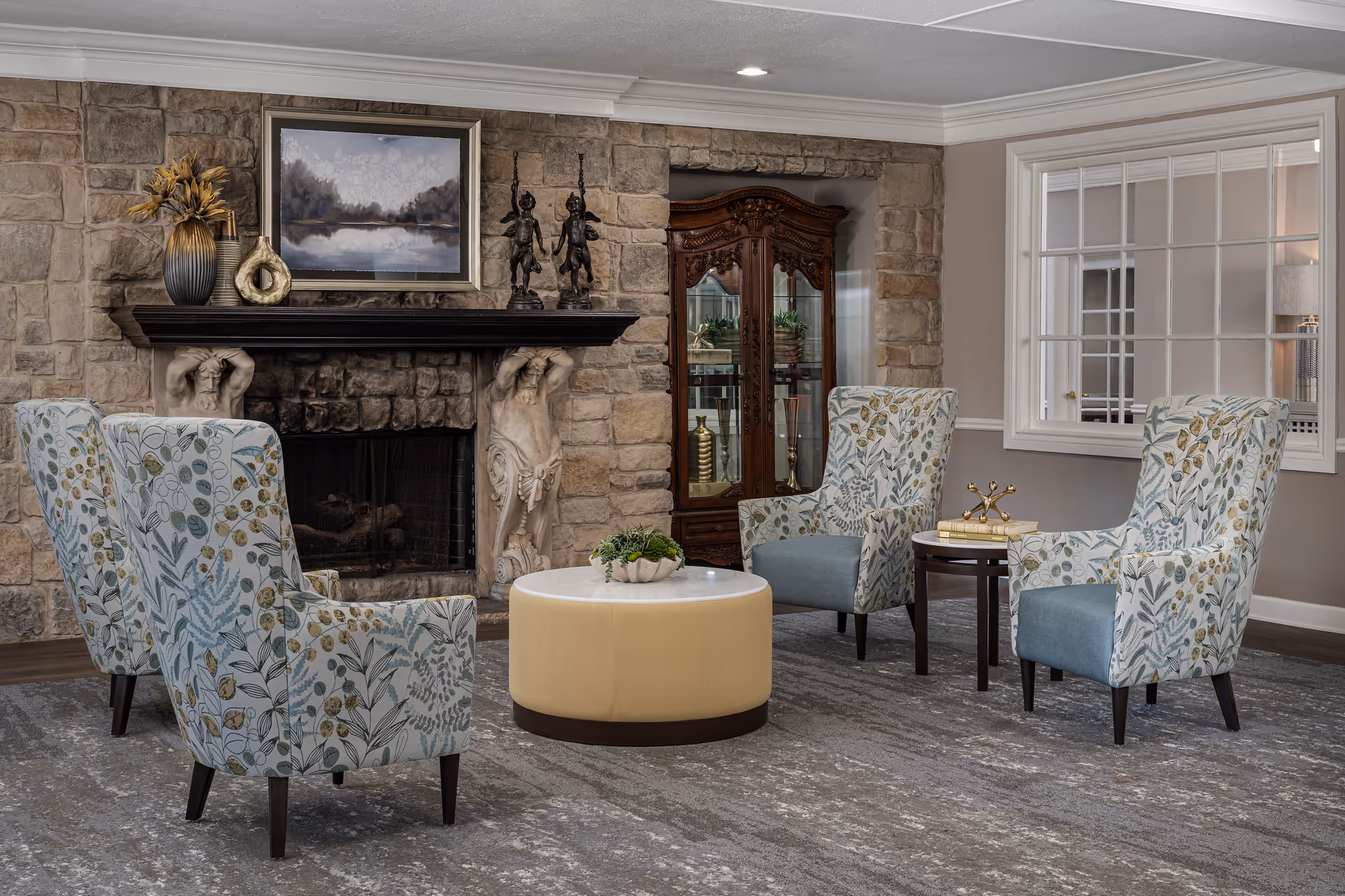A cozy sitting area with four patterned armchairs arranged around a round beige ottoman and a small side table with decorative items. The background features a stone fireplace with sculpted figures supporting the mantel, a framed landscape painting above it, and a wooden display cabinet with glass doors. A large window with white trim is on the right wall.