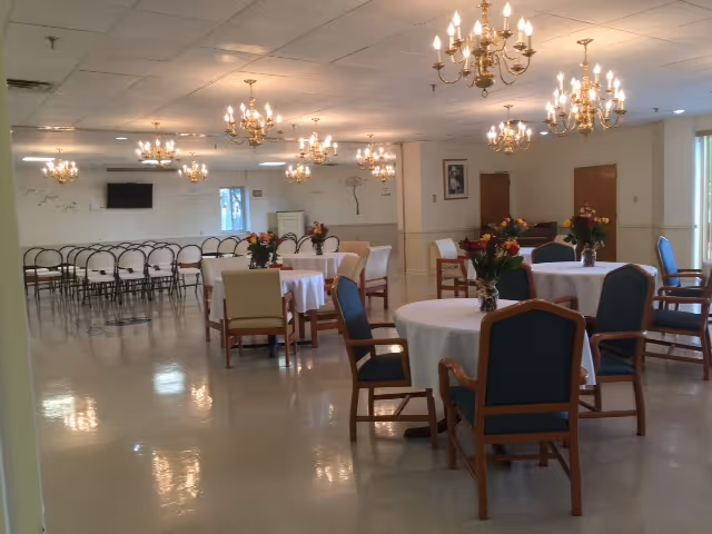 A spacious dining room with round tables covered in white tablecloths, each adorned with a vase of flowers. Several wooden chairs with green and beige cushions surround the tables. The room is well-lit with multiple chandeliers hanging from the ceiling. In the background, there are rows of stacked chairs and a wall-mounted television.