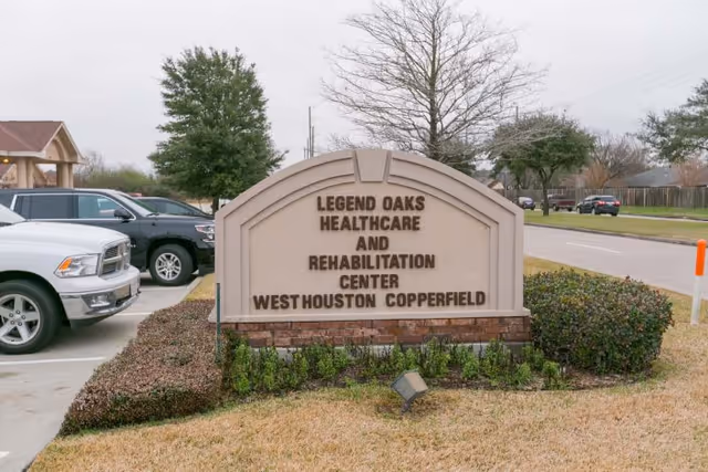 Outdoor view of a stone sign for Legend Oaks Healthcare and Rehabilitation Center West Houston Copperfield, surrounded by trimmed bushes and grass, with parked cars and trees in the background.