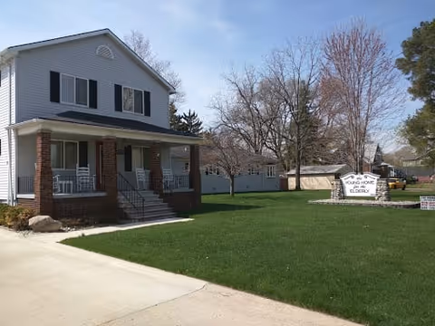 A two-story white building with a front porch and brick pillars, surrounded by a well-maintained green lawn and leafless trees under a clear blue sky. A white sign on the lawn reads 'Young Home For The Elderly.'