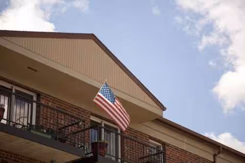 Brick senior living building with balcony and an American flag against a blue sky.