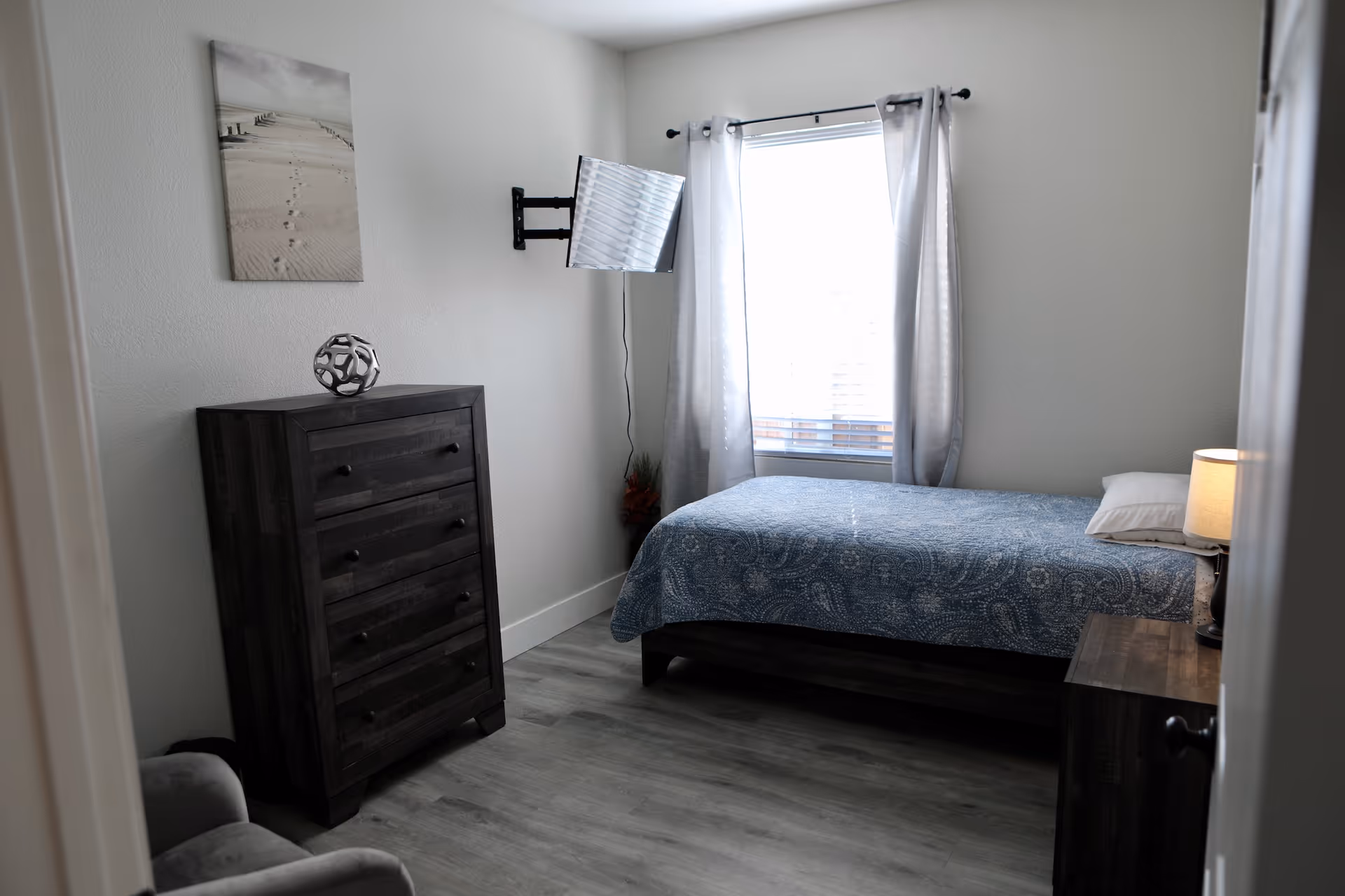 A simple bedroom with a single bed, wooden dresser and nightstand, a wall-mounted TV, and a window with curtains.