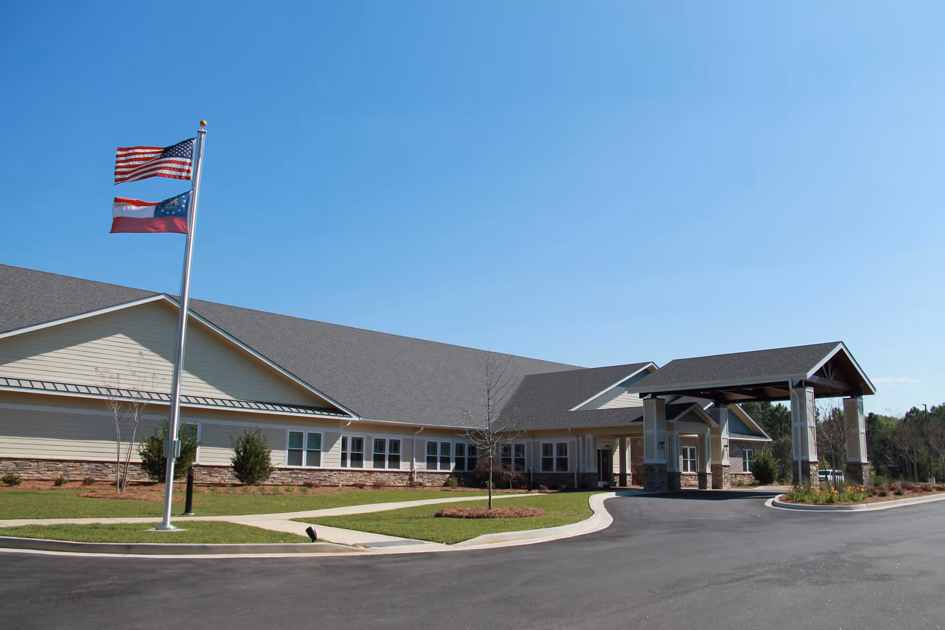 Front entrance of a single-story senior living facility with a covered porte-cochère and flagpoles under a clear blue sky.