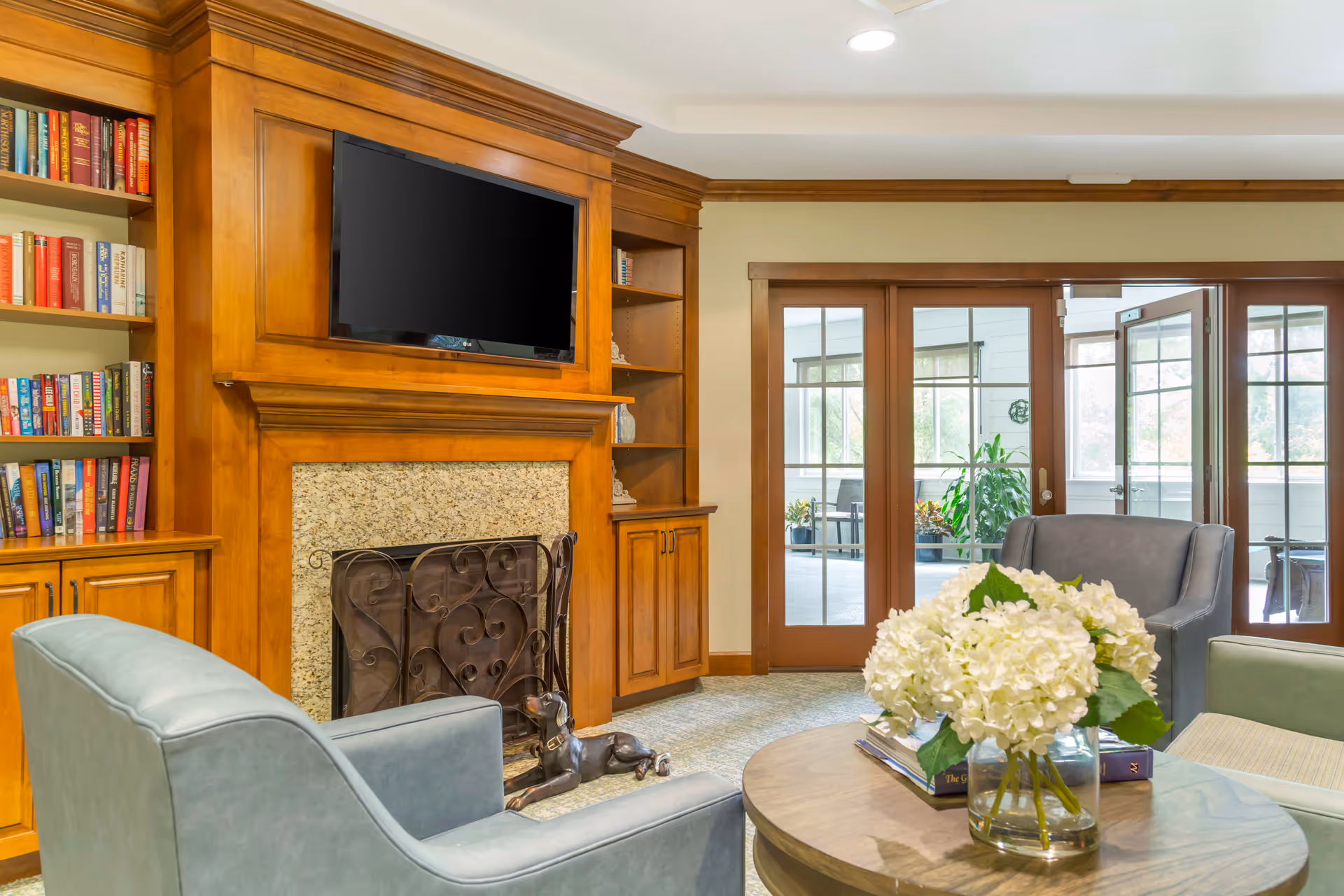 A cozy living room area featuring two blue armchairs and a round wooden coffee table with a glass vase of white flowers. The room has a wooden fireplace with a granite surround and a flat-screen TV mounted above it. Built-in wooden bookshelves filled with books flank the fireplace. Glass doors lead to a bright sunroom with plants and seating visible through the windows.
