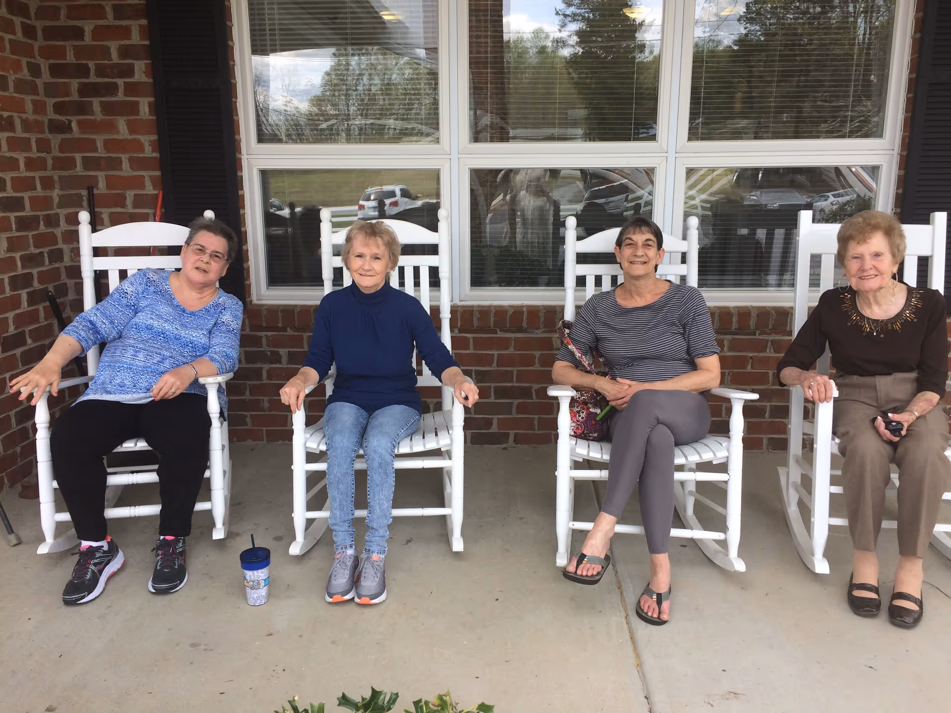Four elderly women sitting on white rocking chairs on a porch in front of a brick wall with large windows, smiling and relaxing.