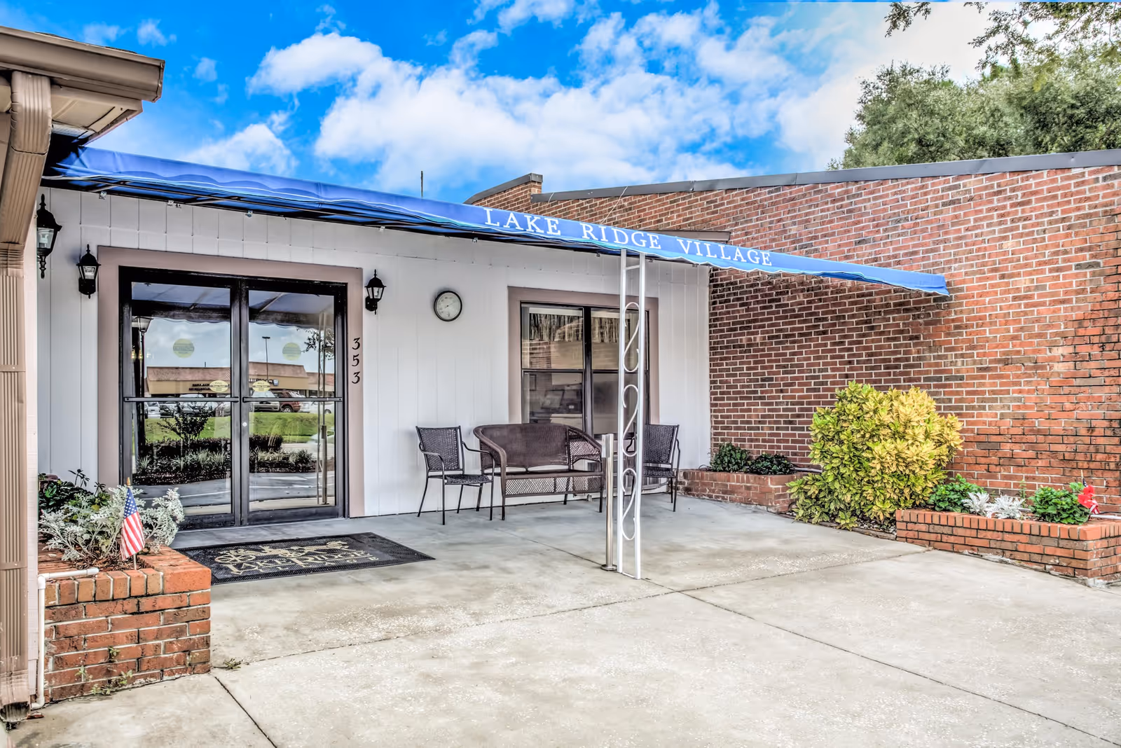 Entrance of Lake Ridge Village with a blue awning over glass double doors, a small seating area, and brick planters.
