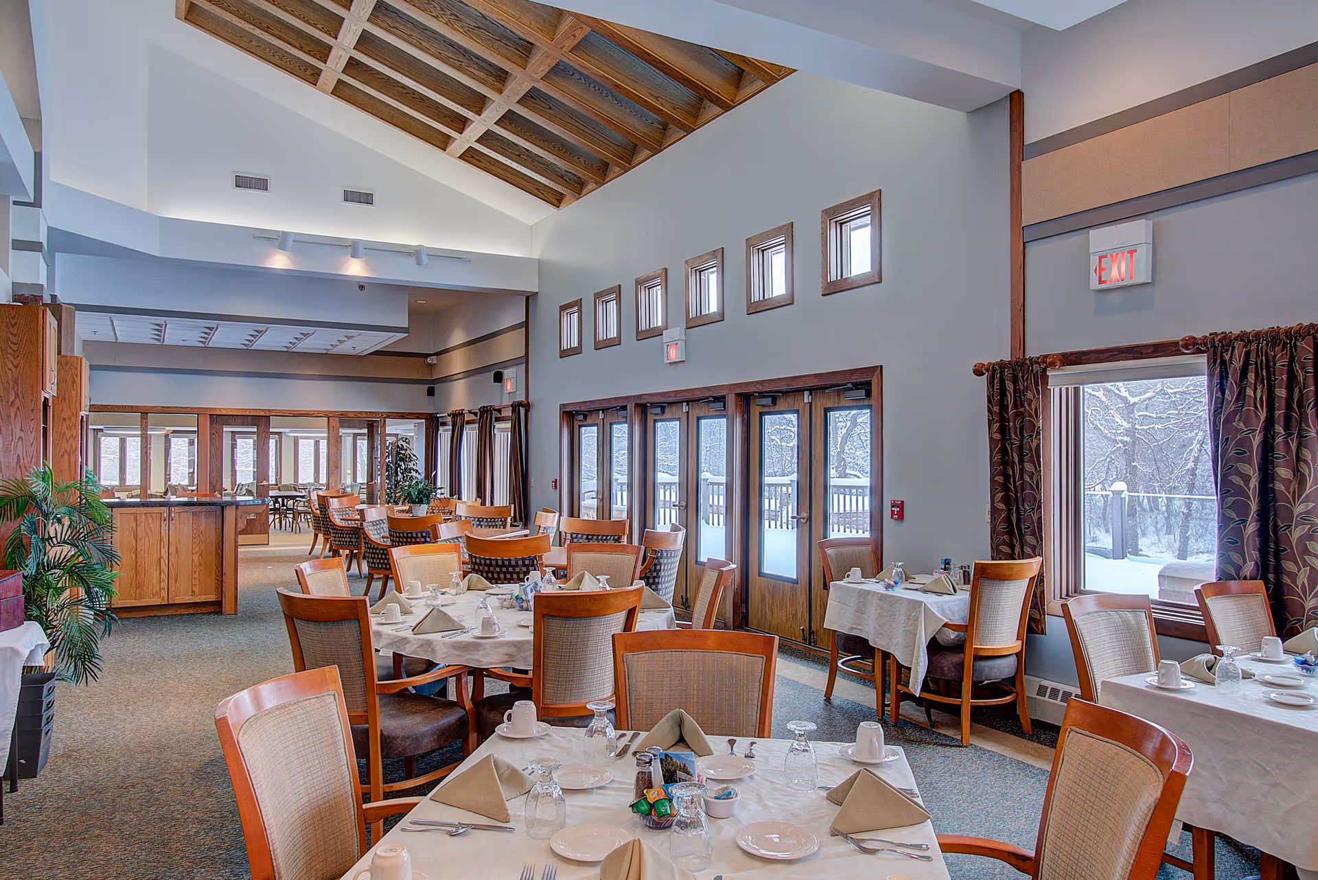 Spacious dining room with set tables and wooden chairs under a vaulted wood-beamed ceiling and large windows.
