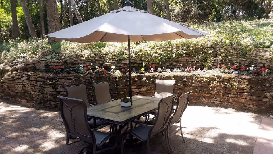 Outdoor patio with a table, six chairs and a large umbrella beside a stone retaining wall and planted flowers.