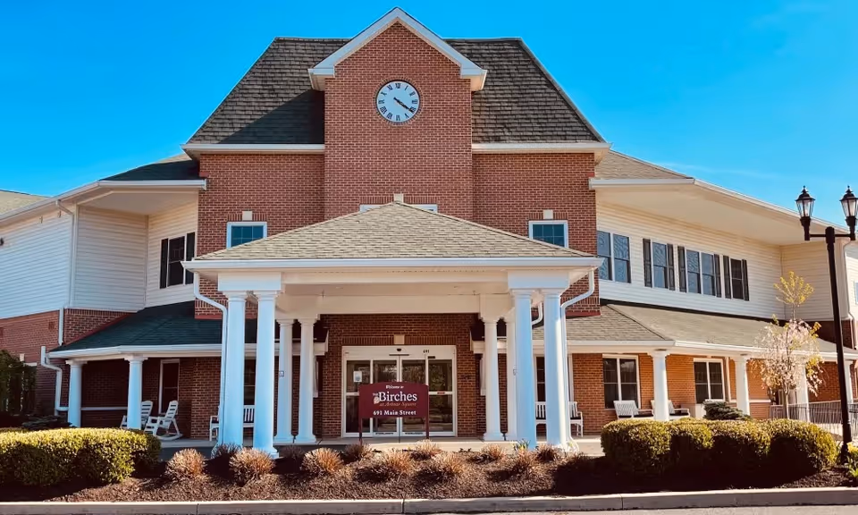 Front entrance of a brick and white senior living building with a columned portico, clock on the facade, and a sign for The Birches.