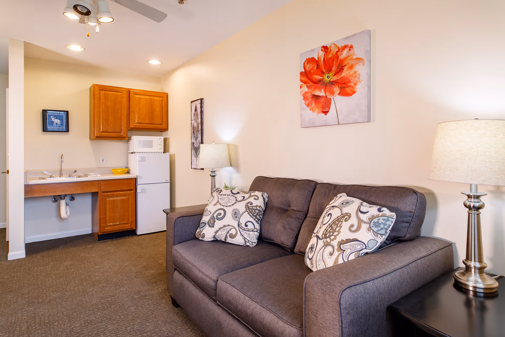 A cozy living area with a dark gray couch adorned with two patterned pillows. Next to the couch is a side table with a lamp. On the wall above the couch is a large painting of a red flower. In the background, there is a small kitchenette with wooden cabinets, a white microwave, and a refrigerator. The room is well-lit with ceiling lights and a ceiling fan.