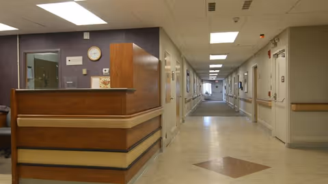 Interior view of a nursing home hallway with a wooden reception desk on the left side and doors lining both sides of the corridor. The hallway is well-lit with ceiling lights and has handrails along the walls.