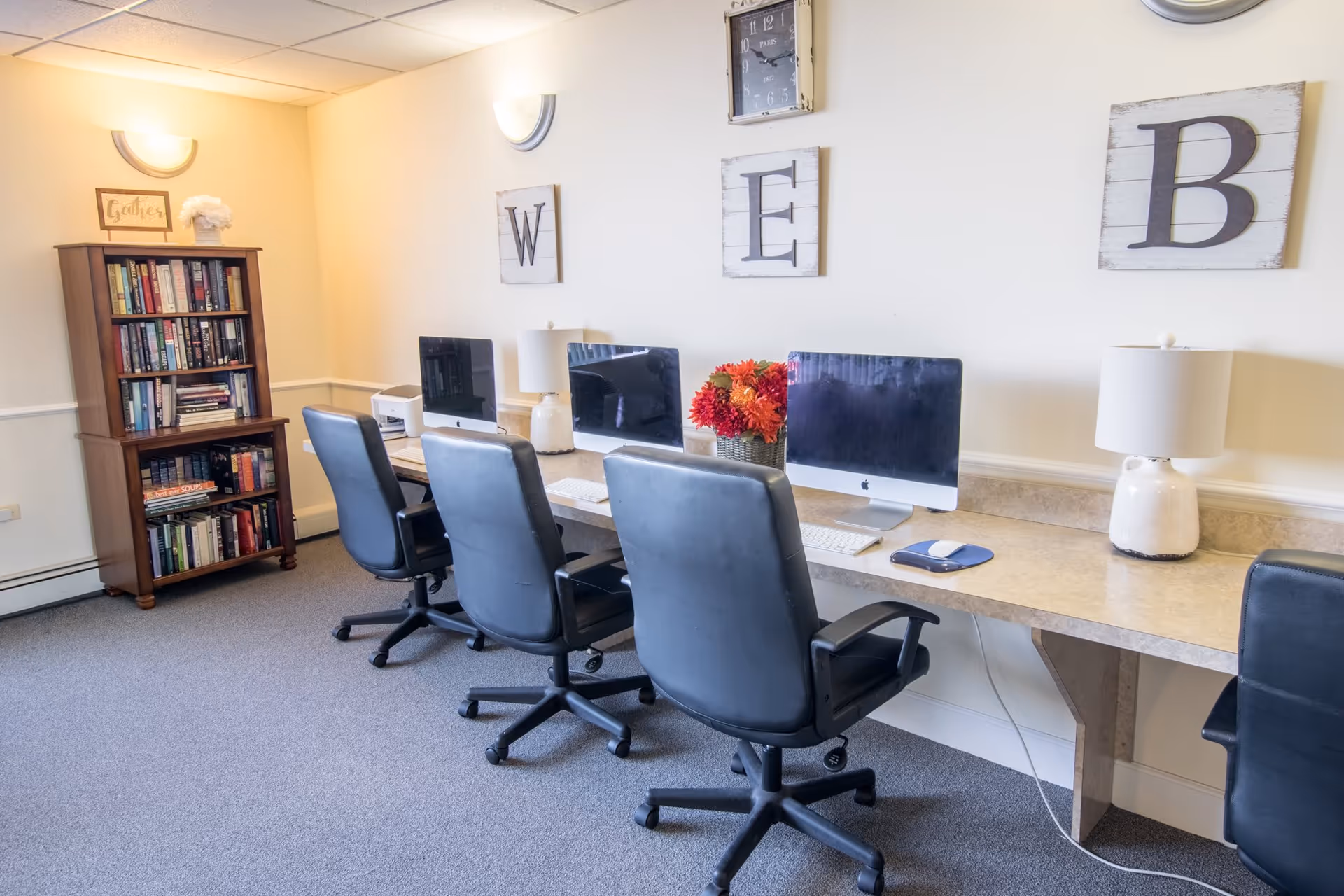 A computer room with four black office chairs in front of a long desk holding three iMac computers, two table lamps, and a vase with red and orange flowers. On the wall above the desk are three wooden plaques with the letters W, E, and B. To the left is a wooden bookshelf filled with books and a small decorative sign on top.