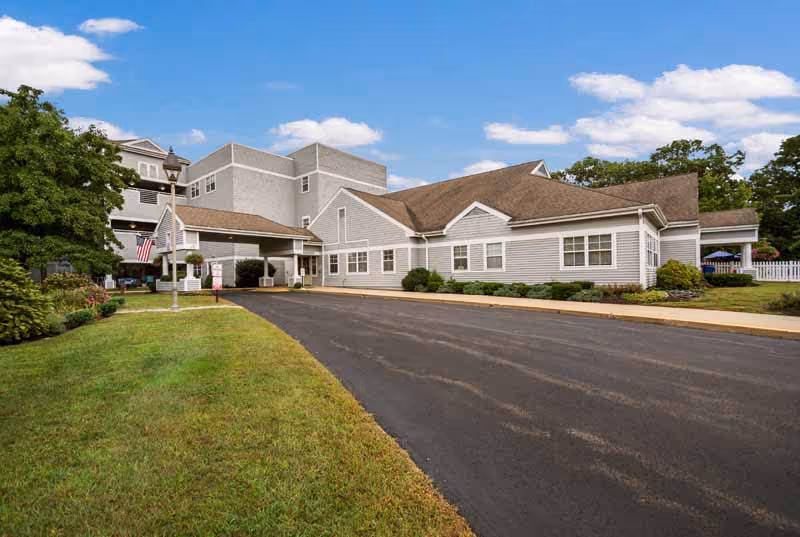 Exterior view of Bride Brook Rehabilitation and Nursing Center showing a multi-section building with gray siding and a brown roof, a paved driveway, green lawn, trees, and a partly cloudy sky.