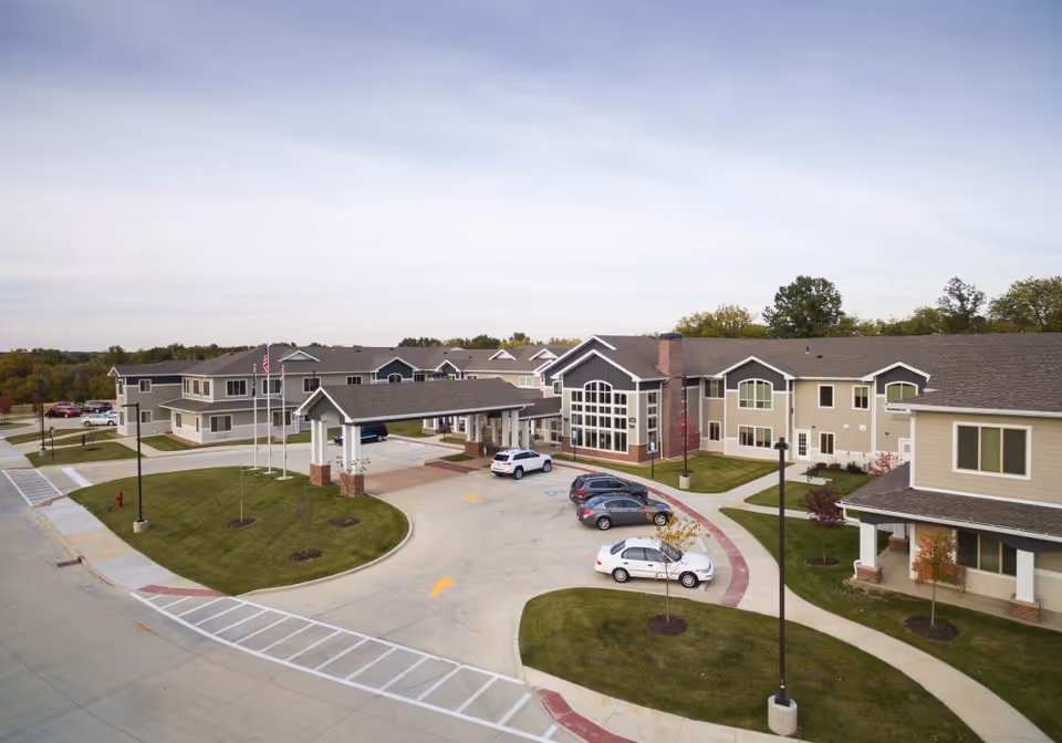 Exterior view of The Landing of O'Fallon senior living facility showing a large two-story building with multiple windows, a covered entrance with a driveway, several parked cars, landscaped green areas with small trees, and a clear sky.