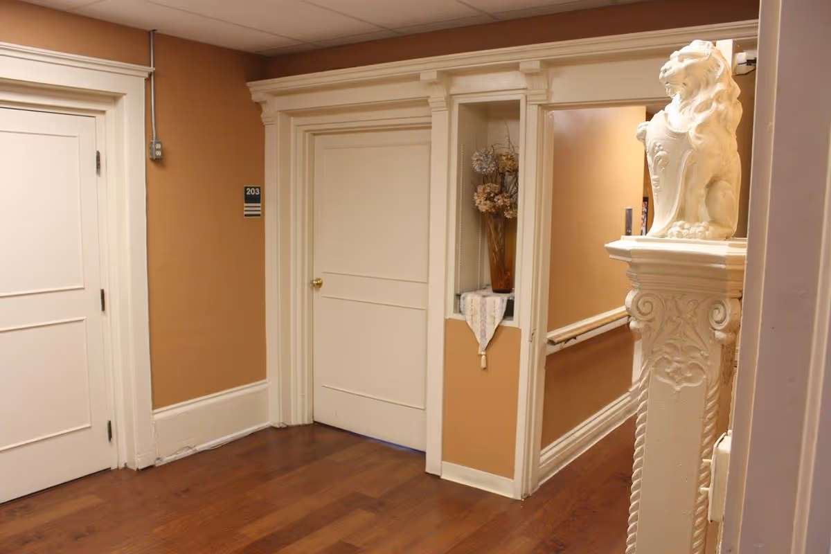 Interior hallway of Singleton Health Care Center with wooden flooring, beige walls, white doors, and decorative molding. A white ornamental lion statue is positioned on a pedestal near the hallway entrance. A small alcove with a vase of dried flowers and a lace cloth is visible on the wall.