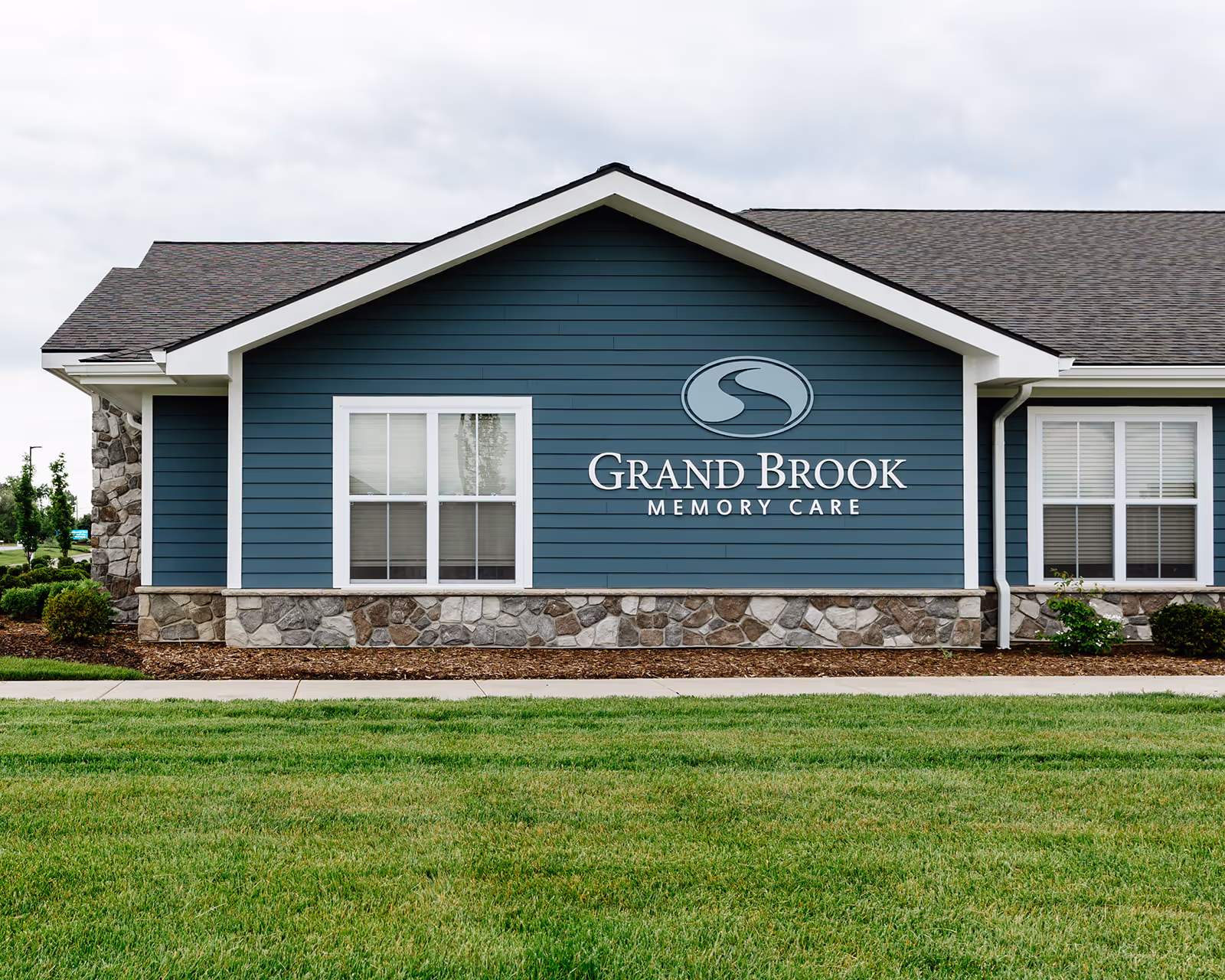 Front exterior of a blue single-story building with stone foundation and a 'Grand Brook Memory Care' sign on the siding.