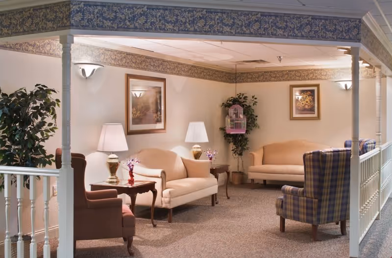A cozy sitting area in a senior living facility with beige sofas, a plaid armchair, wooden side tables with lamps, framed floral artwork on the walls, and potted plants. The space is softly lit and carpeted, with decorative wallpaper border near the ceiling.