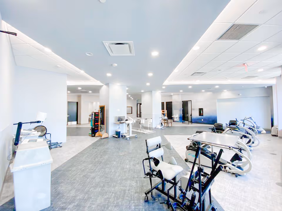 A spacious, well-lit rehabilitation or physical therapy room with various exercise and therapy equipment including stationary bikes and supportive chairs. The room has a modern ceiling with recessed lighting and a mix of light gray and white walls and flooring. Several doors and open areas lead to other parts of the facility.