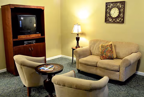 A cozy living room area with a beige loveseat, two beige armchairs, a small round wooden table with magazines, a wooden TV cabinet with an old-style television, a side table with a lamp, and a decorative wall clock.