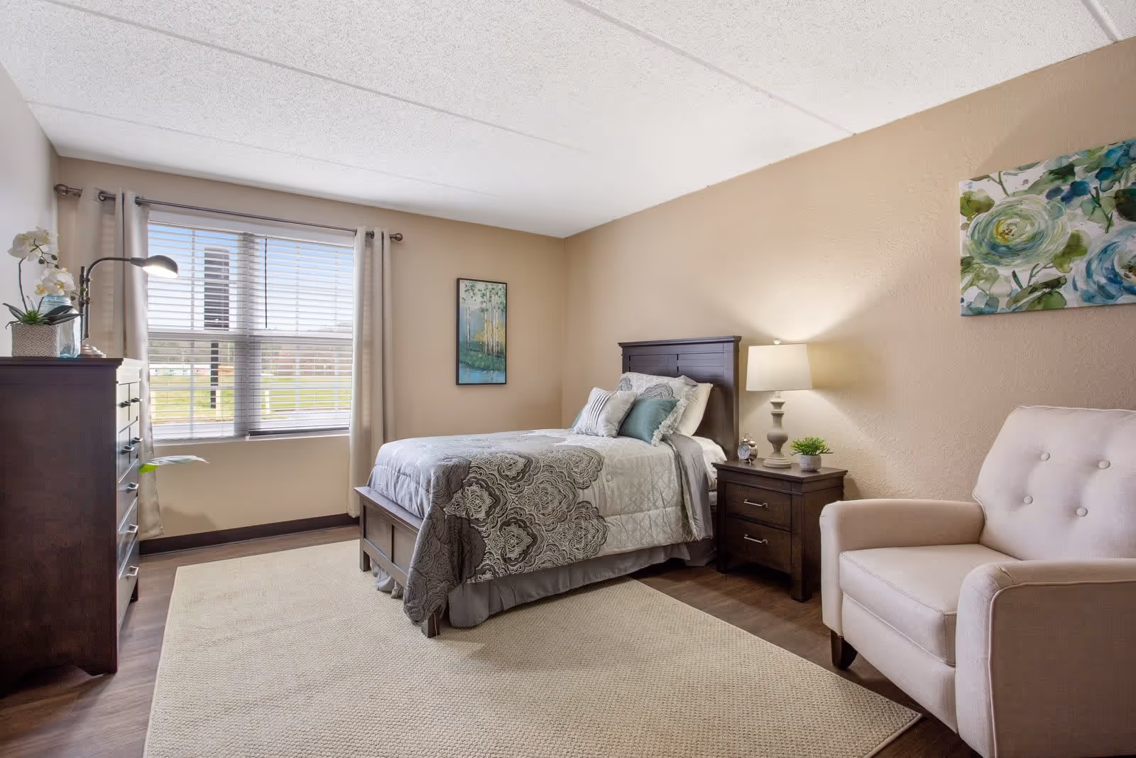 Furnished bedroom with a bed, nightstand and lamp, dresser, window with blinds, and an armchair.