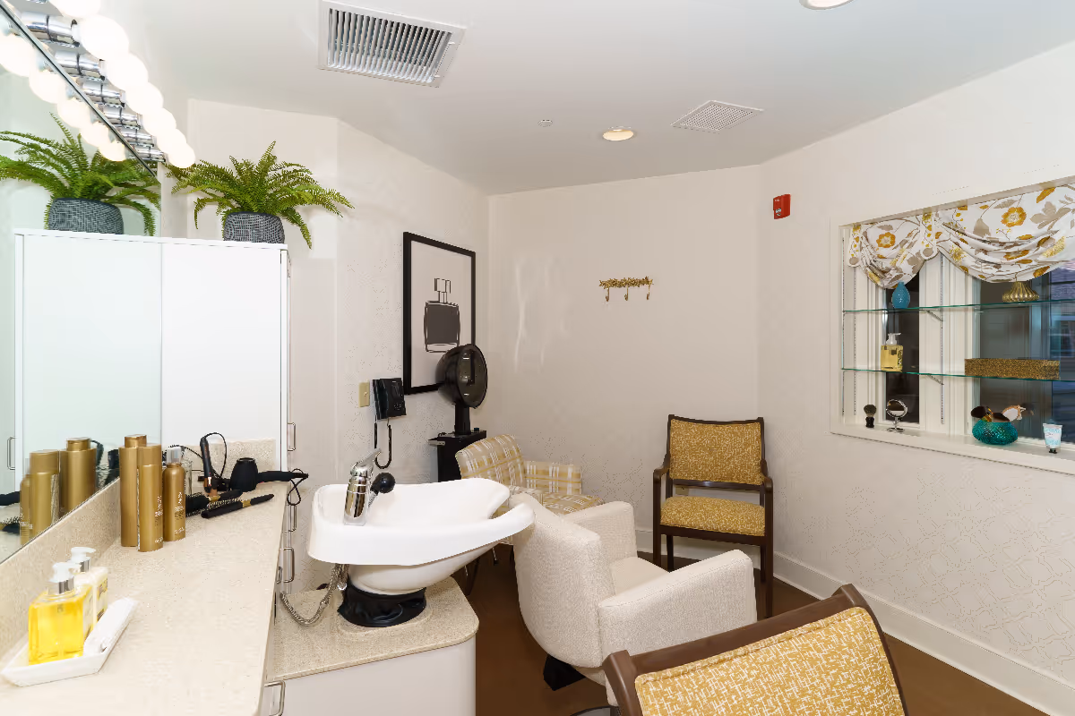 A small salon area in a senior living facility with a white hair washing sink, several chairs including a beige armchair and two yellow patterned chairs, a countertop with hair care products, a large mirror with round lights, a hair dryer, and a window with decorative curtains and shelves displaying small items.