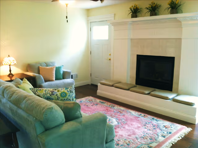 A cozy living room with a light green sofa and armchair, a colorful floral rug on the wooden floor, a white fireplace with a built-in bench topped with cushions, and potted plants on the mantel. A door with a window is visible in the background, along with a floor lamp and a side table with a decorative lamp.