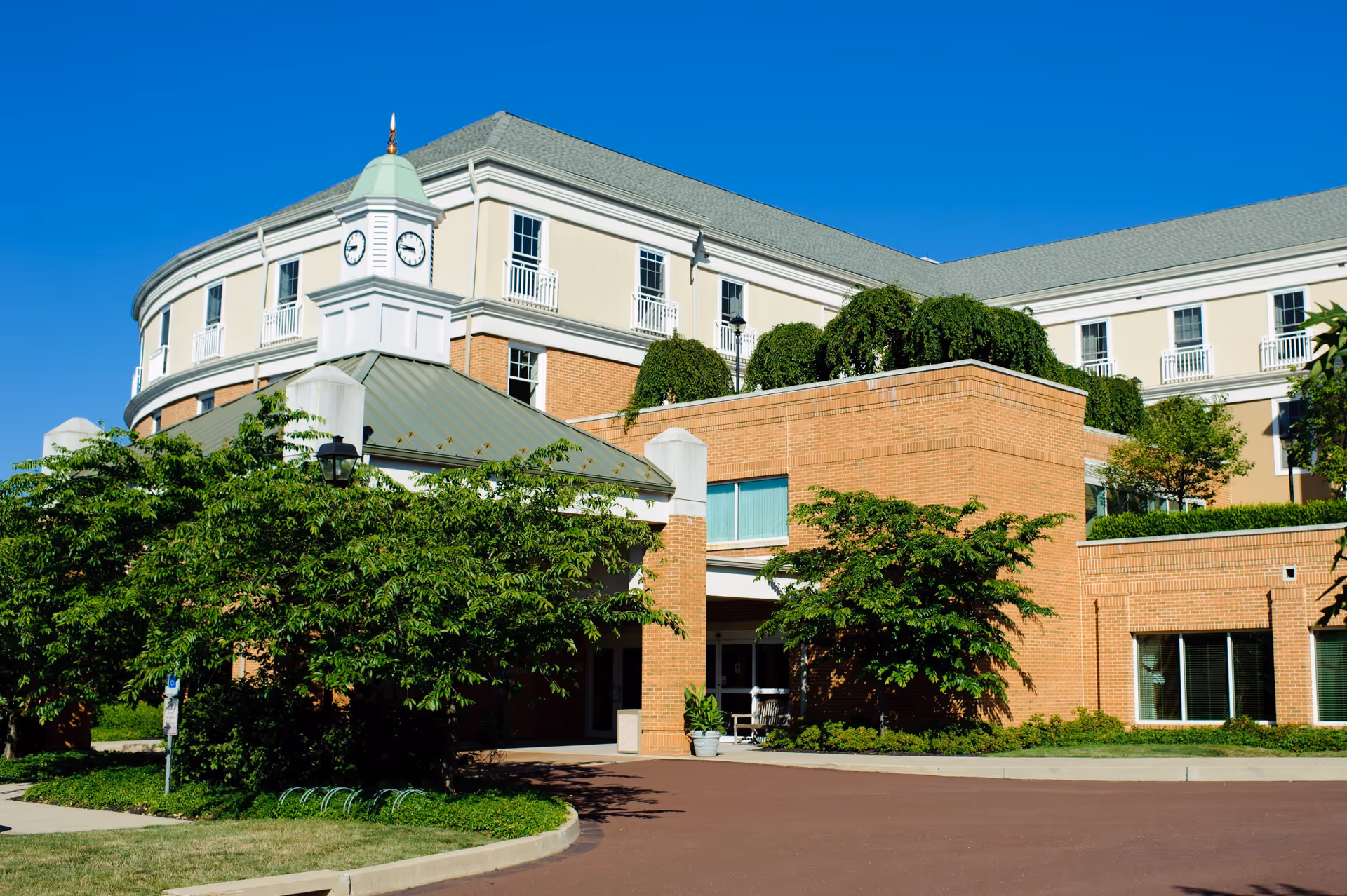 Exterior view of Souderton Mennonite Homes, a CCRC building featuring a clock tower, multiple windows, and lush greenery around the entrance under a clear blue sky.