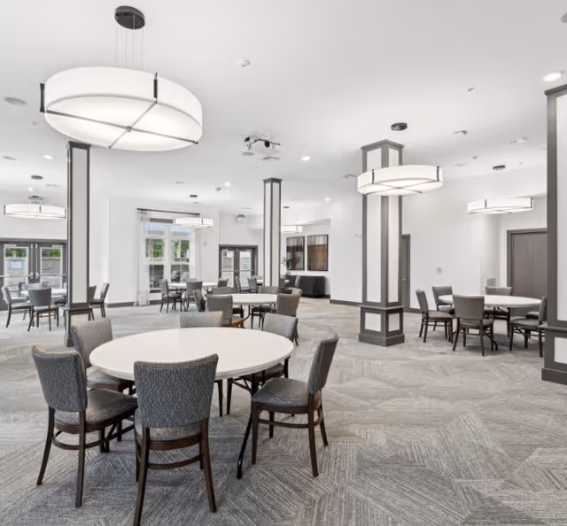 Bright modern communal dining room with round tables, upholstered chairs, tall pillars, and large pendant lights.