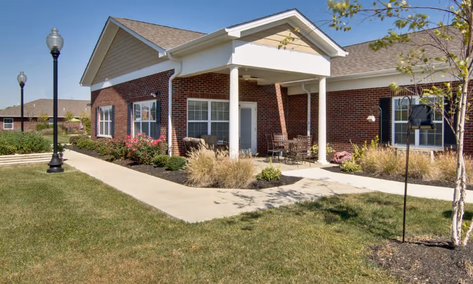 Exterior view of a single-story brick building with white trim and a covered porch area furnished with outdoor chairs and a table. The building is surrounded by a well-maintained lawn, shrubs, and small trees, with a paved walkway and street lamps visible.