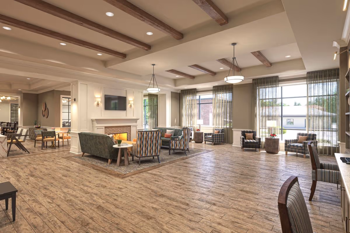 A spacious and well-lit living room area in a senior living facility with wooden flooring, exposed ceiling beams, and large windows with sheer curtains. The room features a fireplace with a mounted TV above it, several armchairs and sofas arranged around small tables, and pendant lights hanging from the ceiling. In the background, there is a dining area with tables and chairs.