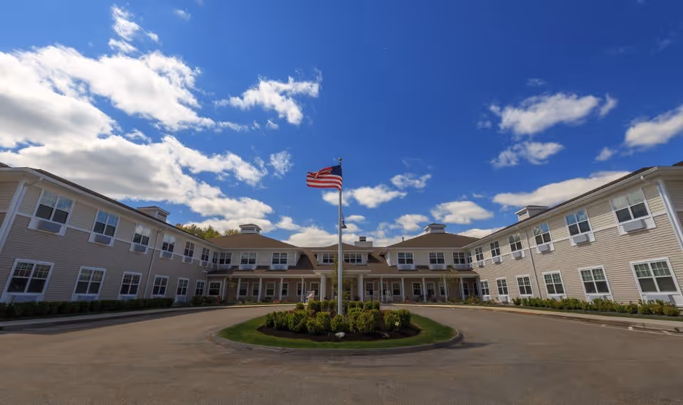 Wide exterior view of a two-story assisted living facility building with beige siding and white trim, featuring many windows and a central entrance. An American flag flies on a flagpole in the middle of a circular driveway with landscaped greenery under a partly cloudy blue sky.