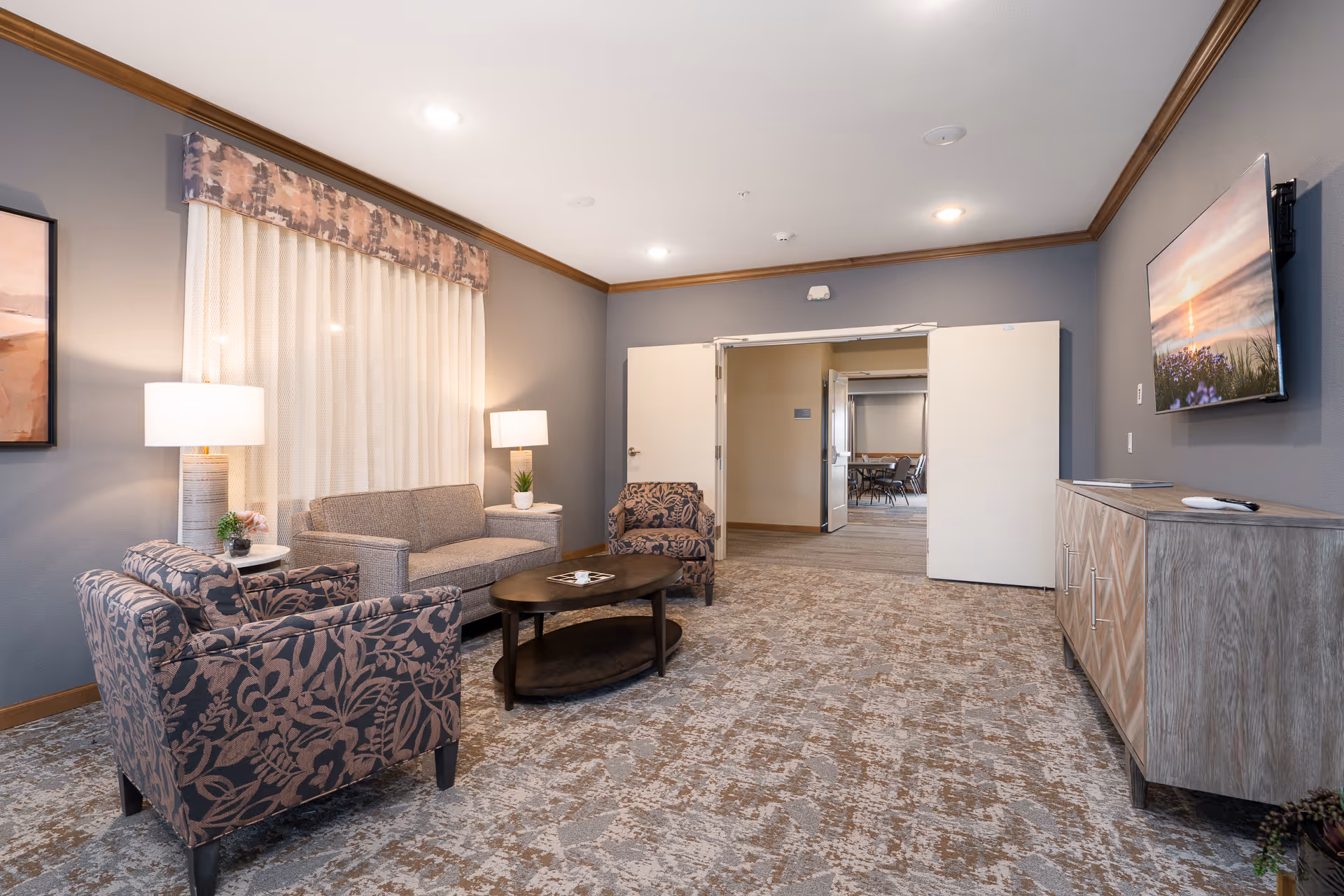 A cozy senior living common area with two patterned armchairs, a gray sofa, a wooden coffee table, two table lamps on side tables, a large window with sheer curtains and a valance, a wall-mounted TV displaying a sunset scene, and a wooden cabinet beneath the TV. The room has gray walls with wood trim and an open doorway leading to another room with tables and chairs.