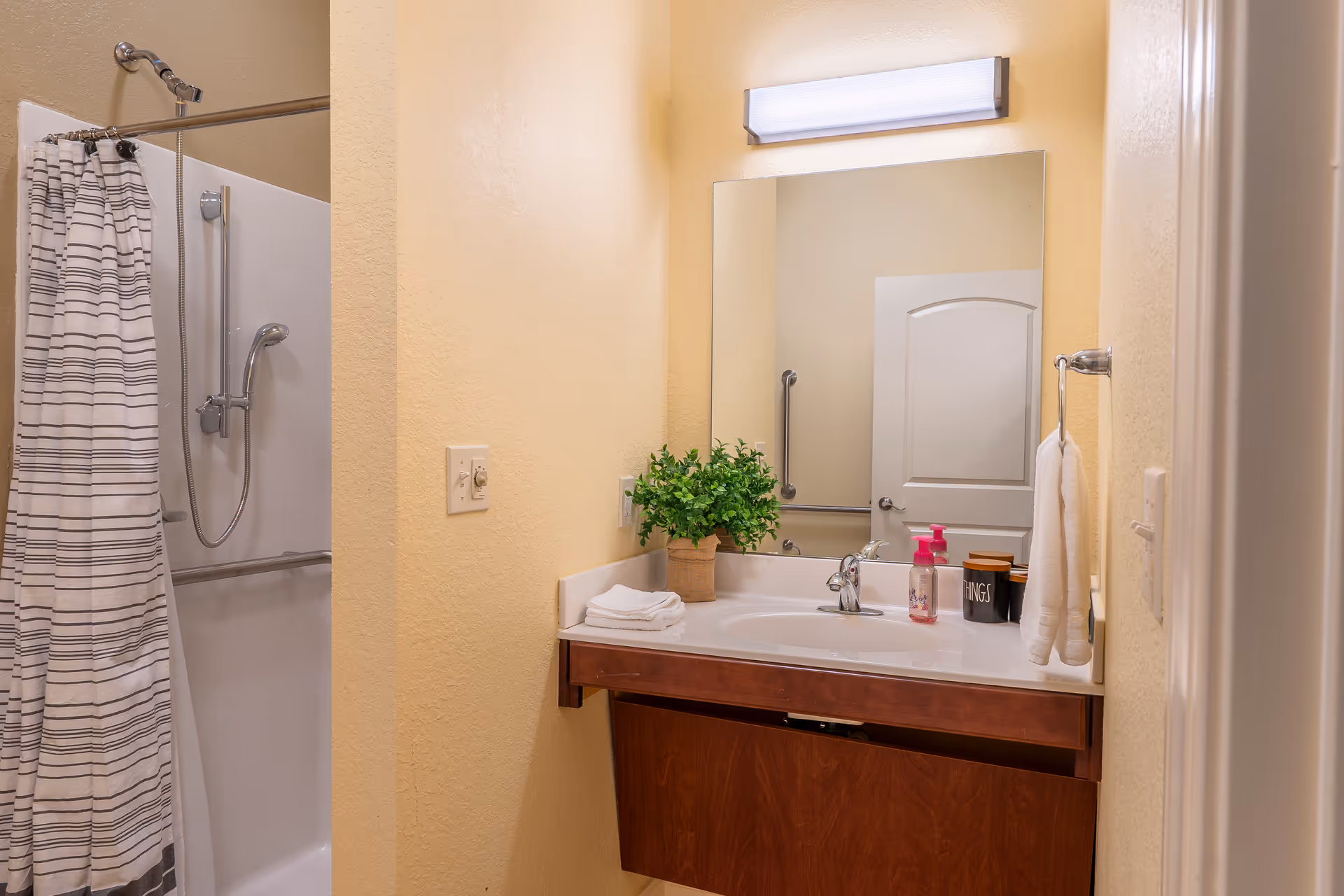 Well-lit bathroom with a sink vanity and mirror, towel and potted plant on the counter, and a shower with a striped curtain.