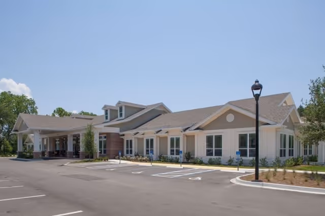 Exterior view of a single-story senior living facility building with a covered entrance, multiple windows, and a parking lot in front under a clear blue sky.