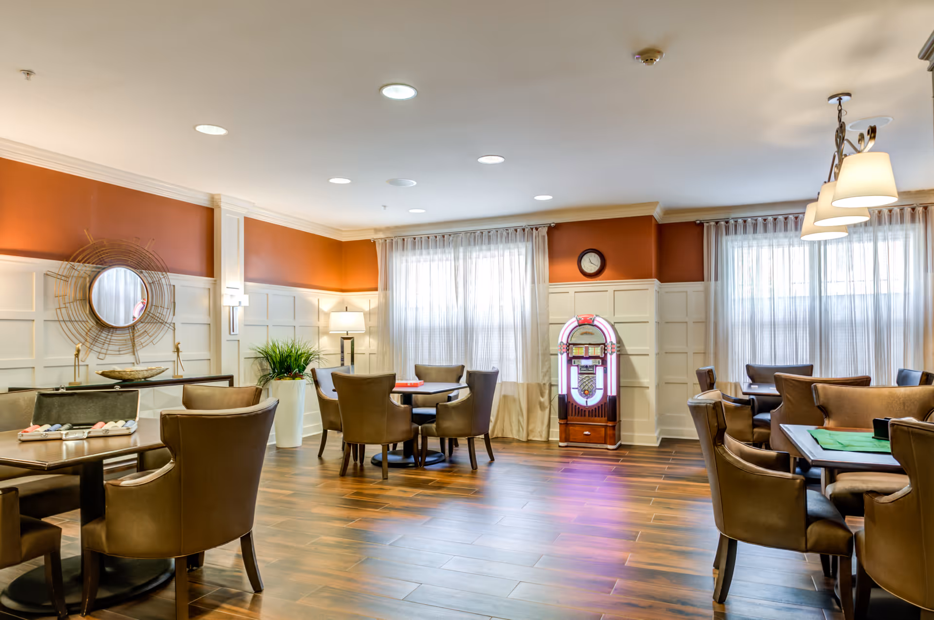 A well-lit common area with several tables and chairs arranged for group activities or dining. The room features wood flooring, white paneled walls with an orange accent wall, large windows with sheer curtains, a decorative round mirror, a potted plant, a standing lamp, a wall clock, and a vintage-style jukebox.