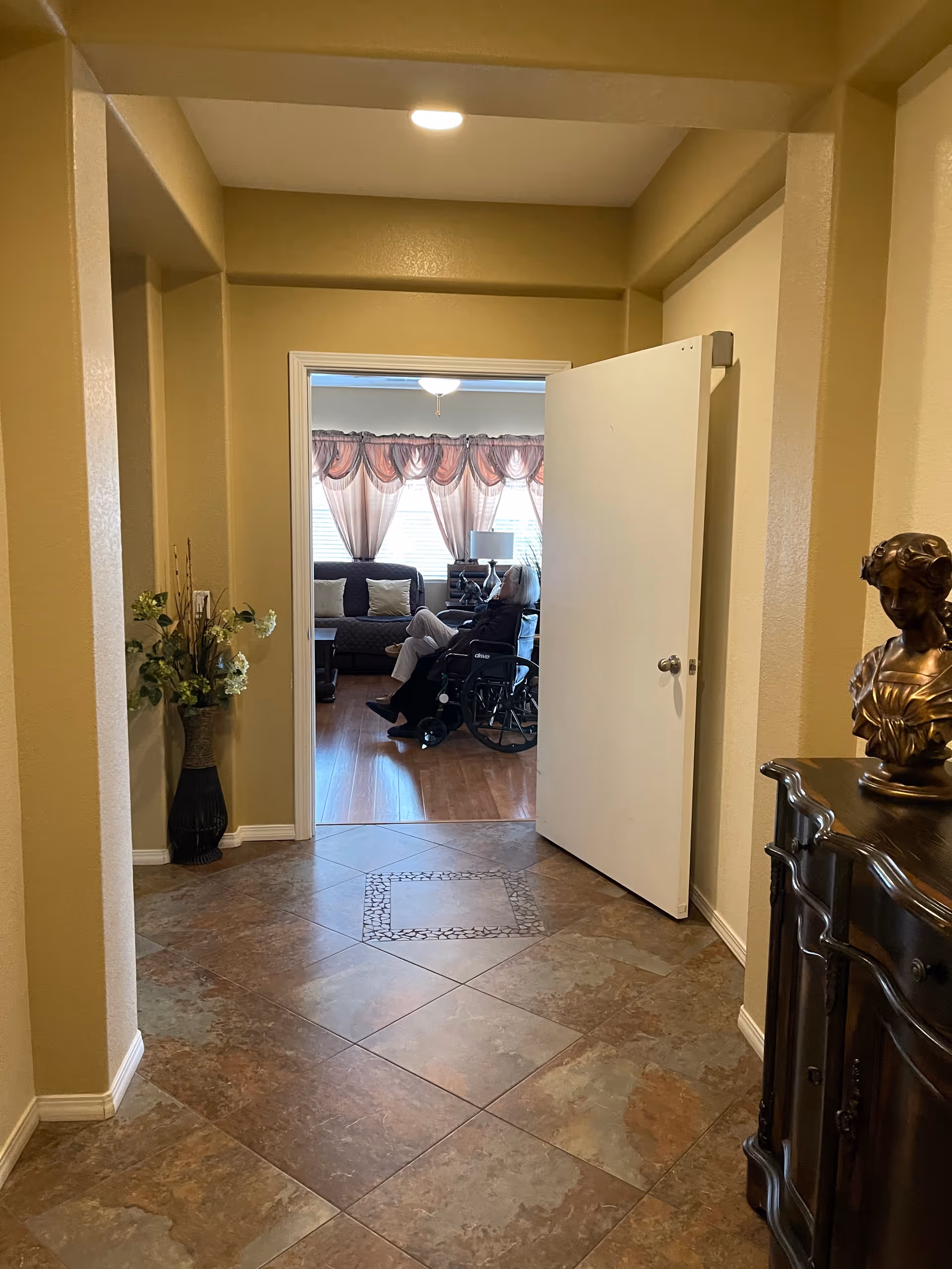 View down a tiled hallway with beige walls leading to a living room where an elderly person in a wheelchair is sitting. The living room has wooden flooring, a dark sofa with cushions, a side table with a lamp, and windows with sheer pink curtains. A decorative vase with flowers is on the left side of the hallway, and a dark wooden cabinet with a bust sculpture is on the right.