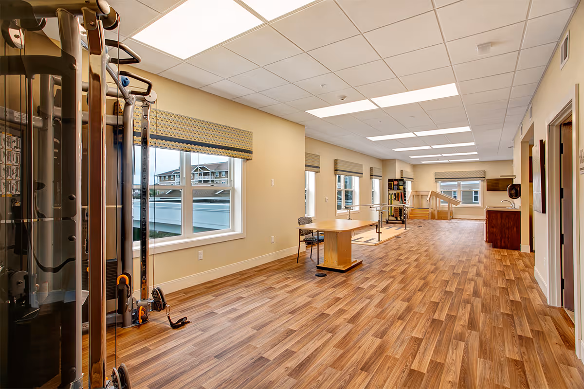 Bright rehabilitation/exercise room with wood floors, windows, exercise equipment and therapy tables.